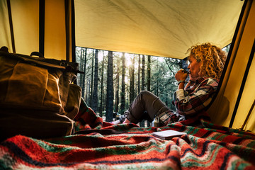 Independent beautiful curly blonde hair woman sit down outside a tent in free wild camping in the forest enjoying a cup of tea and thinking - outdoor natural lifestyle and alternative way of travel