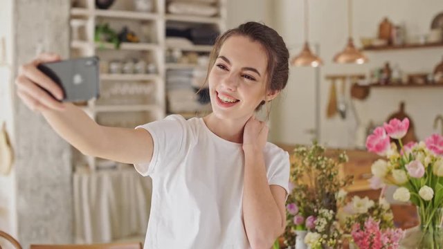 Attractive young brunette woman in white t-shirt taking a selfie photo on smartphone standing at the dining room