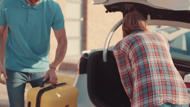 Beautiful traditional family packing for a summer vacation. Happy parents loading suitcases into a car trunk with help of their cute kids in the street.