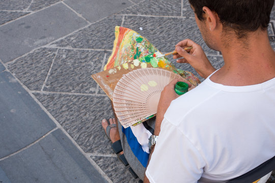 An artist in a Sorrento street mixes paint on his palette as he begins to hand paint a cooling fan.Italy - Image