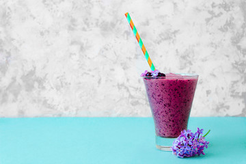 Still life of purple blueberry smoothie with stripped straw and a branch of lilac in glass on white textured background. Healthy vegan lifestyle concept.