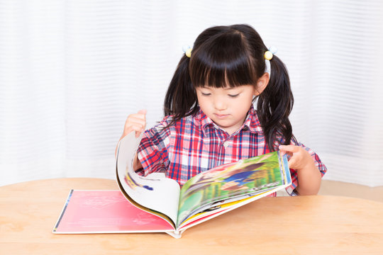 白背景の前で真剣に絵本を読む幼い女の子。幼児、教育、読書、学習、成長、育児イメージ