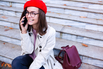 Positive female model in elegant clothes enjoying autumn day, wearing beige jacket and jeans. Outdoor photo of young curly woman in red beret talking on phone and sitting on the stairs.