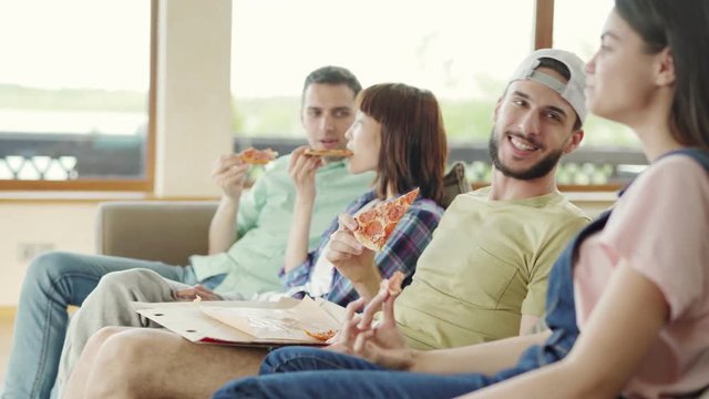 Side view tracking shot of four diverse young friends sitting on sofa, eating delicious pizza and talking. Focus on charismatic man flirting with girlfriend