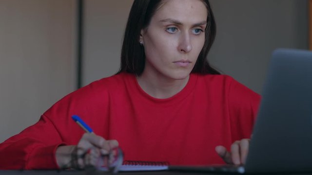 Concept of home education. Cute woman with red sweatshirt overwrites in notepad information from the laptop.