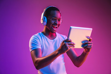 Young african-american man's listening to music in headphones, using tablet in neon light on gradient background. Concept of human emotions, facial expression, summer holidays or weekend, hobby, - obrazy, fototapety, plakaty