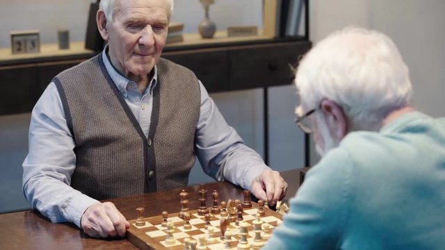 senior man talking while playing chess with friend and pressing button on chess clock at table