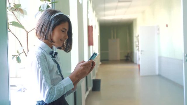 schoolgirl education with smartphones at school concept. Young girl and using smartphone standing by the window in the corridor of the school. correspondent in the messenger, wrote a message. internet