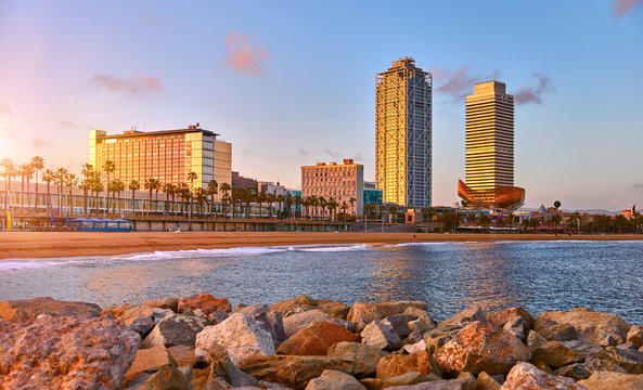 Barcelona, Spain. Panoramic view at Barceloneta beach from sea. View with stones. Sunset landscape with blue sky. Sandy coastline with palms and waves of surf. Popular touristic vacation destination.