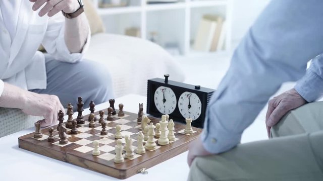 partial view of two senior men playing chess and pressing button on chess clock at table