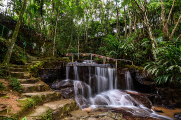 beautiful waterfall in a cave of a sanctuary, with stone stairs and a fence with dozens of colorful ribbons containing promises, surrounded by rainforest - 280234716