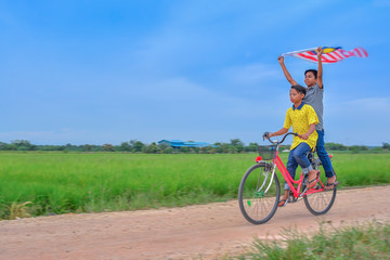 Fototapeta na wymiar independence Day concept - Two happy young local boy riding old bicycle at paddy field holding a Malaysian flag