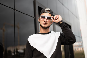 Obraz premium Modern young hipster man with a beard in a stylish black cap in a black-white sweatshirt stands and straightens sunglasses near a gray wall outdoors. Urban guy model resting in the city. Street style.