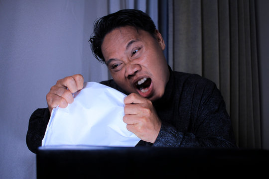 portrait of young Asian man depressed in front of laptop while squeezing and tearing paper, the concept of angry workers