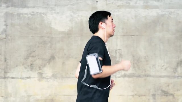 Side view of young determined athletic Asian man wearing headphone and armband with smart phone inside. He is running and jogging on street under big bridge in city in sunny day