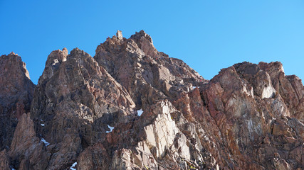 Rocky mountain top. You can see little people. Climbers climb to the top. Large rocks on the sides. Sometimes there is snow. Blue clear sky.