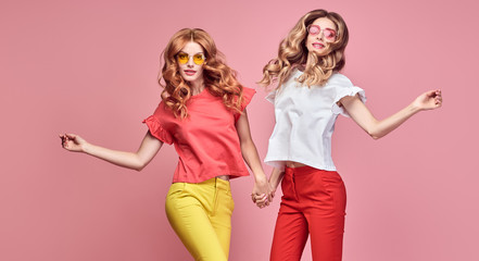 Two fashionable girl jump Smiling in colorful outfit on pink. Beautiful easy-going woman in red yellow pants, Stylish curly hair having fun. Joyful funny slim sisters friends, happy fashion concept