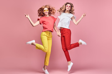 Fototapeta na wymiar Two fashionable girl jump Smiling in colorful outfit on pink. Beautiful easy-going woman in red yellow pants, Stylish curly hair having fun. Joyful funny slim sisters friends, happy fashion concept