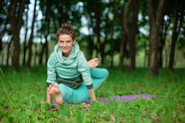 Thin brunette girl plays sports and performs beautiful and sophisticated yoga poses in a summer park. Green lush forest on the background. Woman doing exercises on a yoga mat