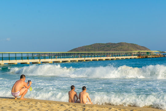 Two Middle-aged Men Of Asian Appearance Are Sitting On The Seashore Nearby And Resting, Meeting The Approaching Big Waves, And The Third Is Taking Pictures Of Them From Behind.