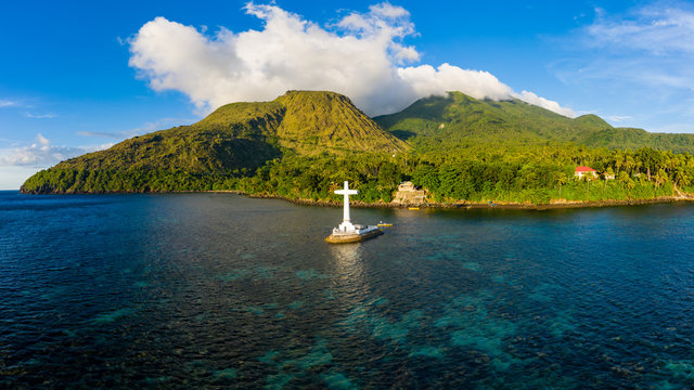 Aerial drone view of a large cross marking a sunken cemetery with evening light on background volcanos on a tropical island (Camiguin, Philippines)