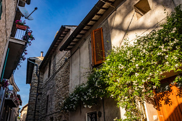 A narrow alley in the medieval village of Bevagna. Perugia, Umbria, Italy. The blue sky between the buildings. The flowering climbing jasmine, on the wall of a house. Stone and brick walls.