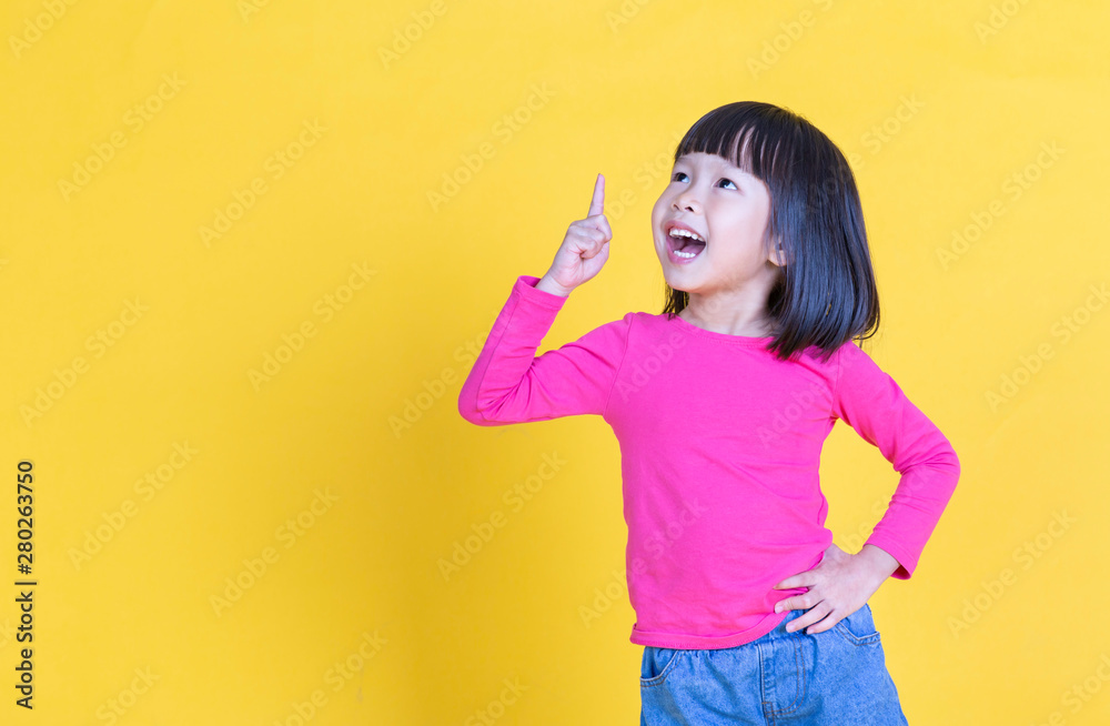 Wall mural portrait of young little asian girl hand point up isolated on yellow background with copy space. edu