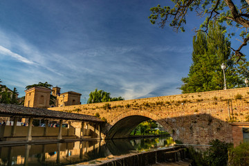 Fototapeta na wymiar The ancient wash-house and the masonry bridge over the river, in the medieval village of Bevagna. Perugia, Umbria, Italy. Blue sky at sunset. Trees and vegetation. The reflection on the water surface.