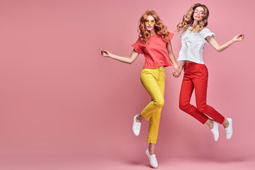 Fototapeta na wymiar Two fashionable girl jump Smiling in colorful outfit on pink. Beautiful easy-going woman in red yellow pants, Stylish curly hair having fun. Joyful funny slim sisters friends, happy fashion concept