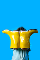 a boy in a light t-shirt covered his face with yellow rubber boots. symbolizes autumn on blue background