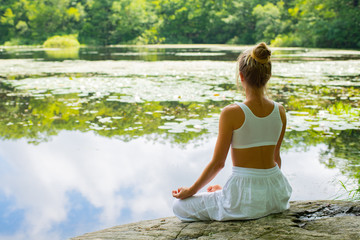 Attractive woman is practicing yoga sitting in lotus pose on stone near lake.