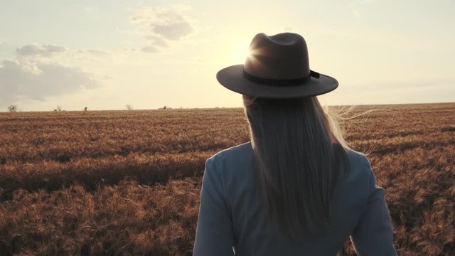 girl in a trendy hat walking across a field of wheat at sunset