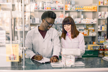 Confident multiethnical Male And Female Pharmacists In Pharmacy. African American man pharmacist making notes on clipboard during inventory in pharmacy