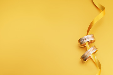 Two wedding rings connected by a ribbon on a yellow background with copy space, top view