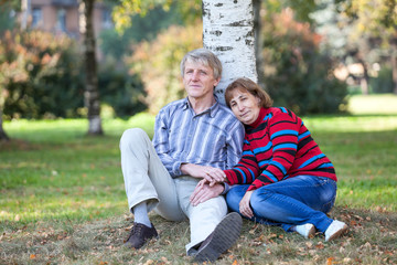Fototapeta premium Senior couple sitting under birch tree on earth in autumn park, holding hands and resting