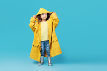 Happy funny child posing on blue studio background. Girl is wearing yellow waterproof raincoat and...