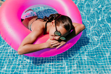 beautiful teenager girl floating on pink donuts in a pool. Wearing sunglasses and smiling. Fun and summer lifestyle