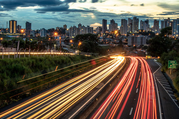 Fototapeta premium Trail of light caused by vehicular traffic in SP-294, Comandante Joao Ribeiro Barros Highway with buildings from downtown in the background, in Marília, during a late afternoon.