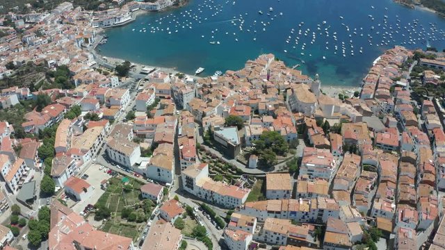 Drone flight over tiled roofs, narrow streets, white houses, church of resort coastal town, boats and yachts on a beautiful bay. Aerial view of Cadaques on background of scenic seascape, Spain