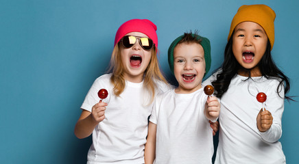 Three joyful friends kids in white t-shirts and colorful hats hold sweet lollipop candies happy smiling hugging