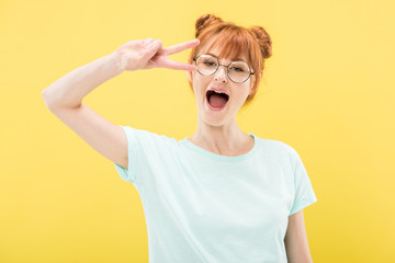 excited redhead girl in glasses and t-shirt showing peace sign isolated on yellow