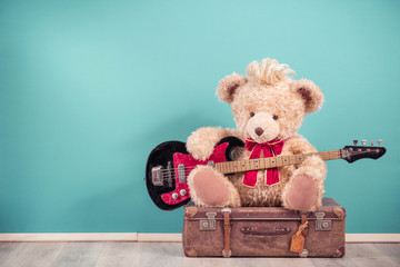 Retro Teddy Bear toy with hairstyle playing bass guitar and sitting on old leather travel luggage....