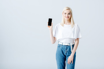 Naklejka premium beautiful happy blonde girl showing smartphone with blank screen isolated on grey