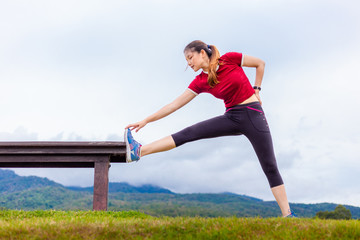 Beautiful young Asian woman stretching during her morning exercise  at a park on a cloudy sky day, with mouintains and sky background, healthy lifestyle, copy space