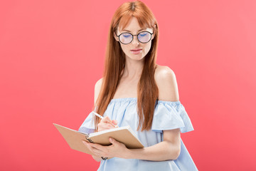 attractive redhead woman in glasses holding pencil and textbook isolated on pink