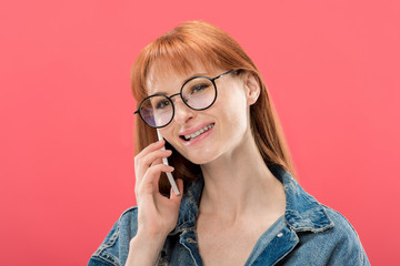attractive redhead girl in glasses and denim jacket talking on smartphone isolated on pink