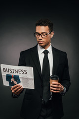 handsome african american businessman in glasses and suit holding coffee to go and reading newspaper on dark background