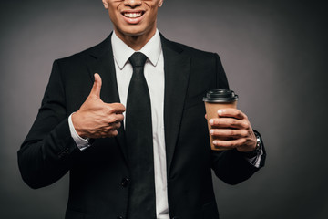 cropped view of smiling african american businessman in suit holding coffee to go and showing thumb up on dark background