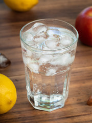 Close up view on glass full of water and melting ice cubes. Yellow lemons, red apple and glass on brown desk. Selective soft focus. Blurred background