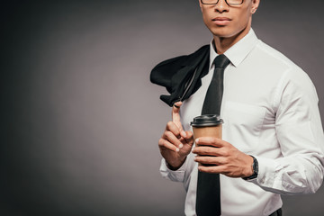 cropped view of african american businessman holding blazer and coffee to go on dark background